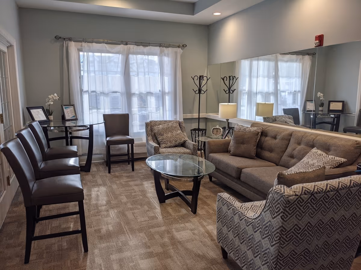 A cozy living room area with a large window covered by sheer white curtains. The room features a gray sofa with patterned and solid pillows, two patterned armchairs, a glass coffee table, a small round glass side table with a lamp and clock, and a coat rack in the corner. There are four brown chairs lined up against the left wall near a small round glass table with framed certificates and a small plant. The walls are painted light gray and a large mirror covers one wall, reflecting the room.