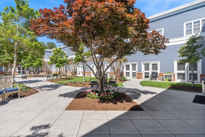 Outdoor courtyard area at Morningside of Wilmington featuring a paved walkway, benches, a central tree with red leaves surrounded by flowers, and a two-story building with blue siding and white trim in the background under a clear blue sky.