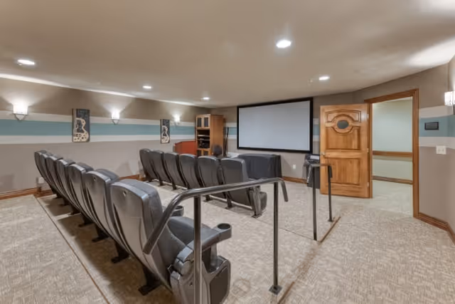 A small theater room with two rows of black cushioned seats facing a large white projection screen. The room has beige carpet, light-colored walls with a blue and white horizontal stripe, wall-mounted lights, and a wooden door open to an adjacent room.
