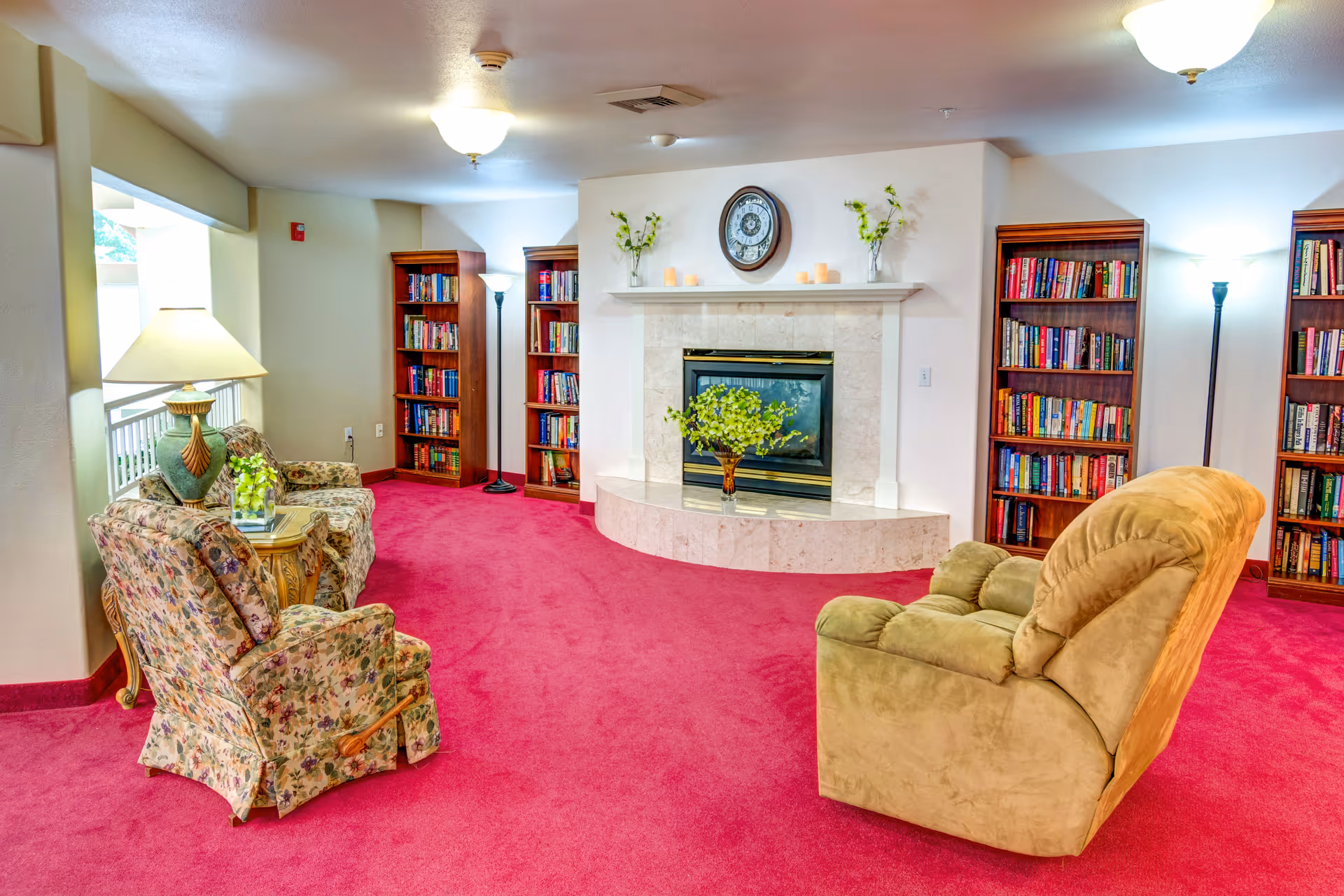 A cozy living room area with a red carpet, a beige recliner chair, and two floral-patterned armchairs around a small wooden table with a lamp and a plant. The room features a white fireplace with a clock and decorative candles on the mantel, flanked by tall wooden bookshelves filled with books. Two floor lamps provide additional lighting.