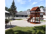 Outdoor playground area with a wooden play structure including slides and climbing features, surrounded by gravel and grass. In the background, there are buildings with gray roofs and white siding, along with some trees.