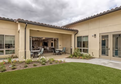 Outdoor patio area of a senior living facility with tables and chairs under a covered section, surrounded by beige walls with windows and doors. There is a small garden with flowers and plants next to a green lawn, under a cloudy sky.