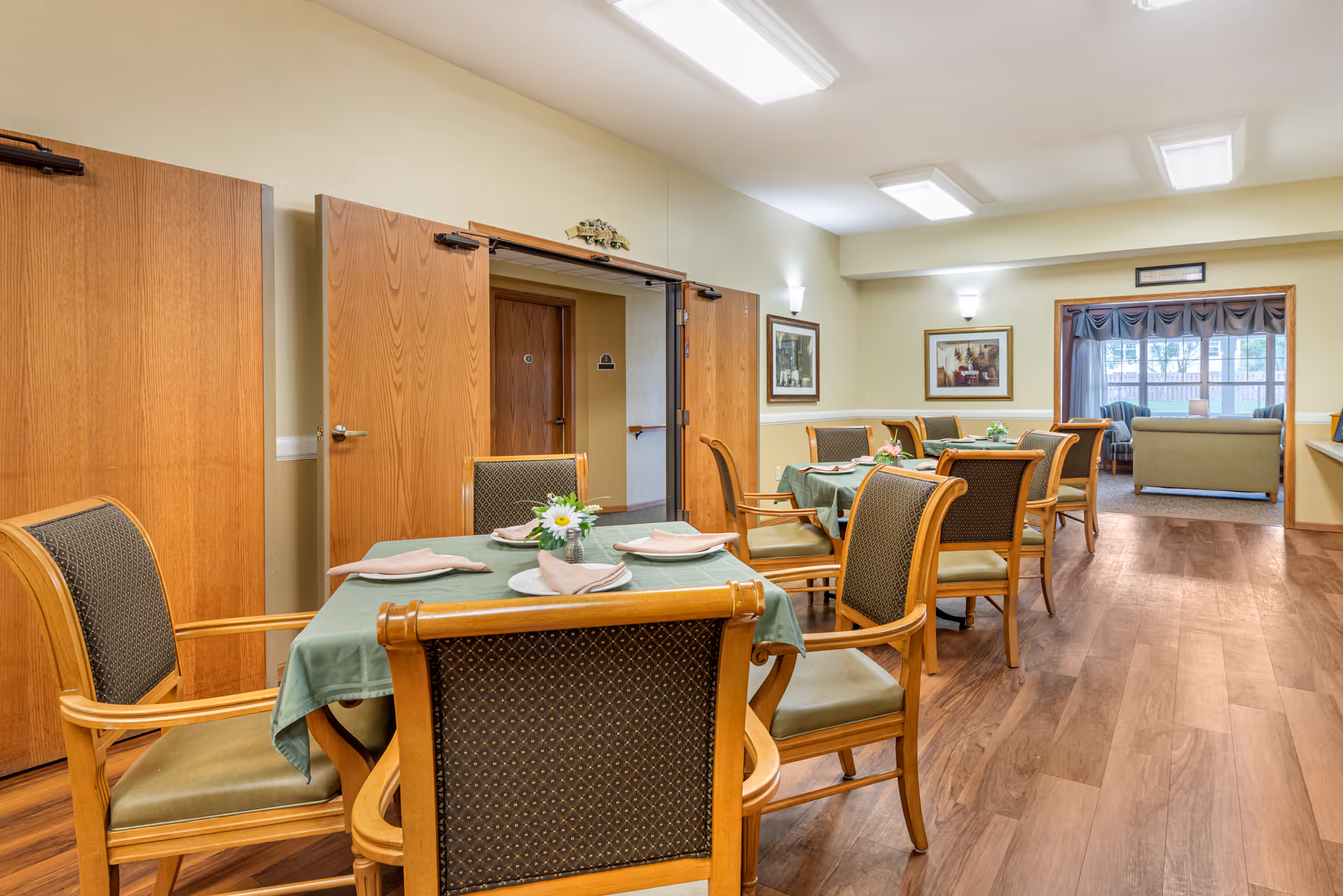 Interior view of a dining area in a senior living facility with wooden tables covered with green tablecloths, each set with napkins and small flower arrangements. Wooden chairs with cushioned seats surround the tables. The room has wood flooring, beige walls, and fluorescent ceiling lights. In the background, there is a sitting area with sofas near a window with curtains.