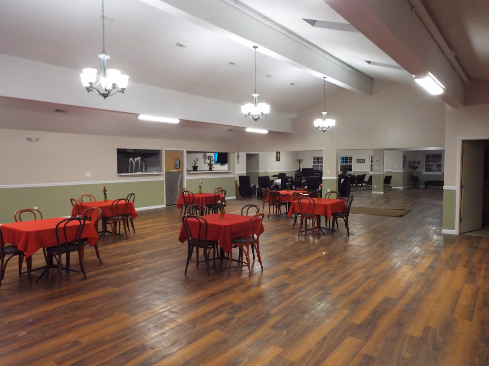 A spacious dining room with several small square tables covered with red tablecloths, each table surrounded by four wooden chairs. The room has wooden flooring, light green and beige walls, and multiple ceiling lights including chandeliers. In the background, there are additional seating areas with black chairs and a door leading to another room.