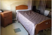 A small bedroom in a nursing facility with a single bed covered by a patterned quilt, a wooden nightstand with a telephone on it, and a window with curtains partially drawn.