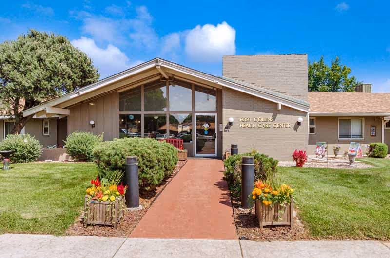 Front exterior view of a single-story health care center building with a peaked roof, large glass entrance doors, and a sign that reads 'Fort Collins Health Care Center'. The building is surrounded by green grass, bushes, and flower planters along a red walkway leading to the entrance. The sky is blue with a few clouds.