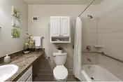 A clean and modern bathroom featuring a white toilet, a bathtub with a shower curtain, a countertop with a sink, and a wall-mounted cabinet above the toilet. The walls are light-colored, and there are decorative plants and towels adding a touch of greenery and comfort.