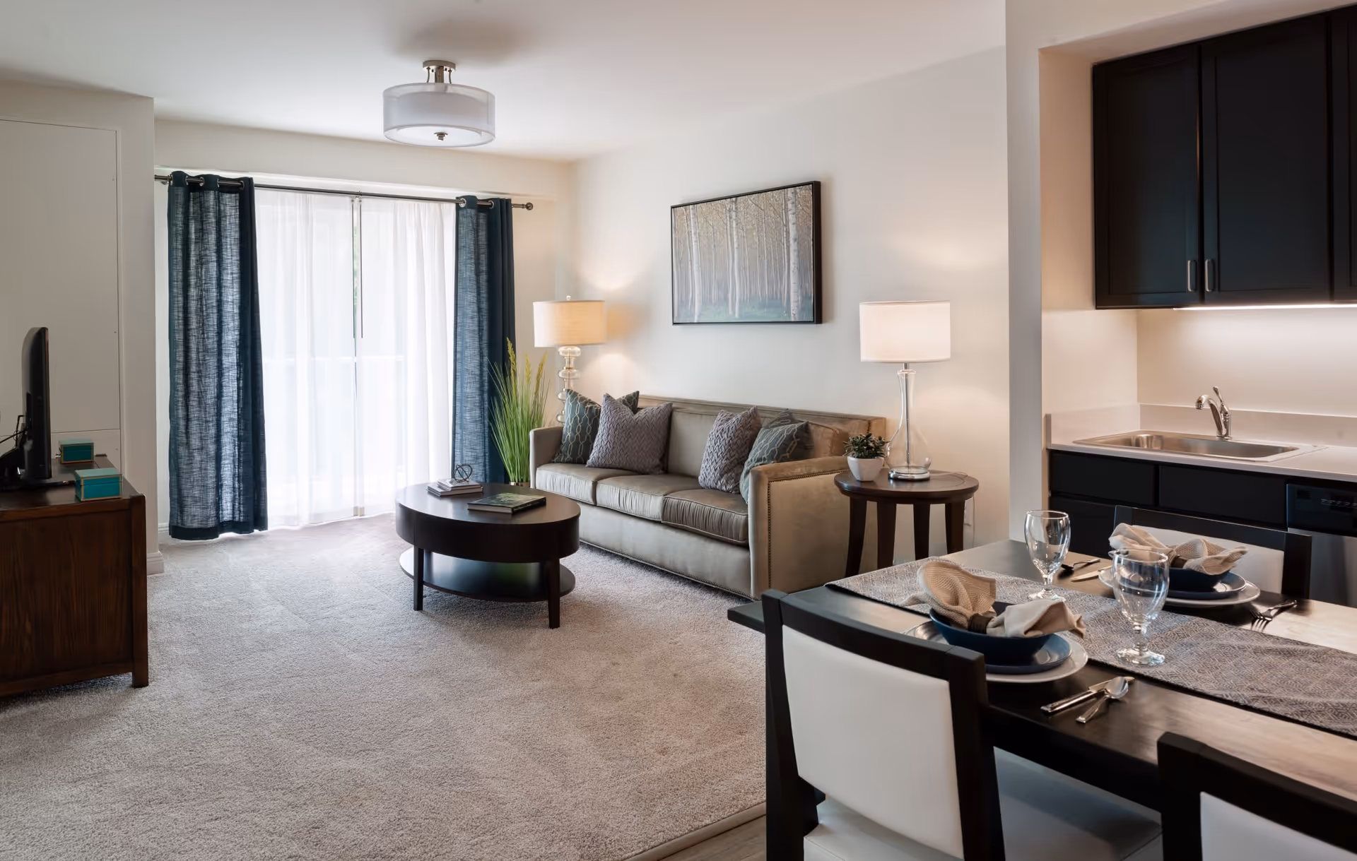 A bright living room and kitchen area in a senior living facility. The living room features a beige sofa with several cushions, two side tables with lamps, a round coffee table, and a TV on a wooden stand. Large windows with sheer white curtains and dark blue drapes allow natural light to fill the room. The kitchen area includes dark cabinets, a sink, and a dining table set for two with plates, glasses, and napkins.
