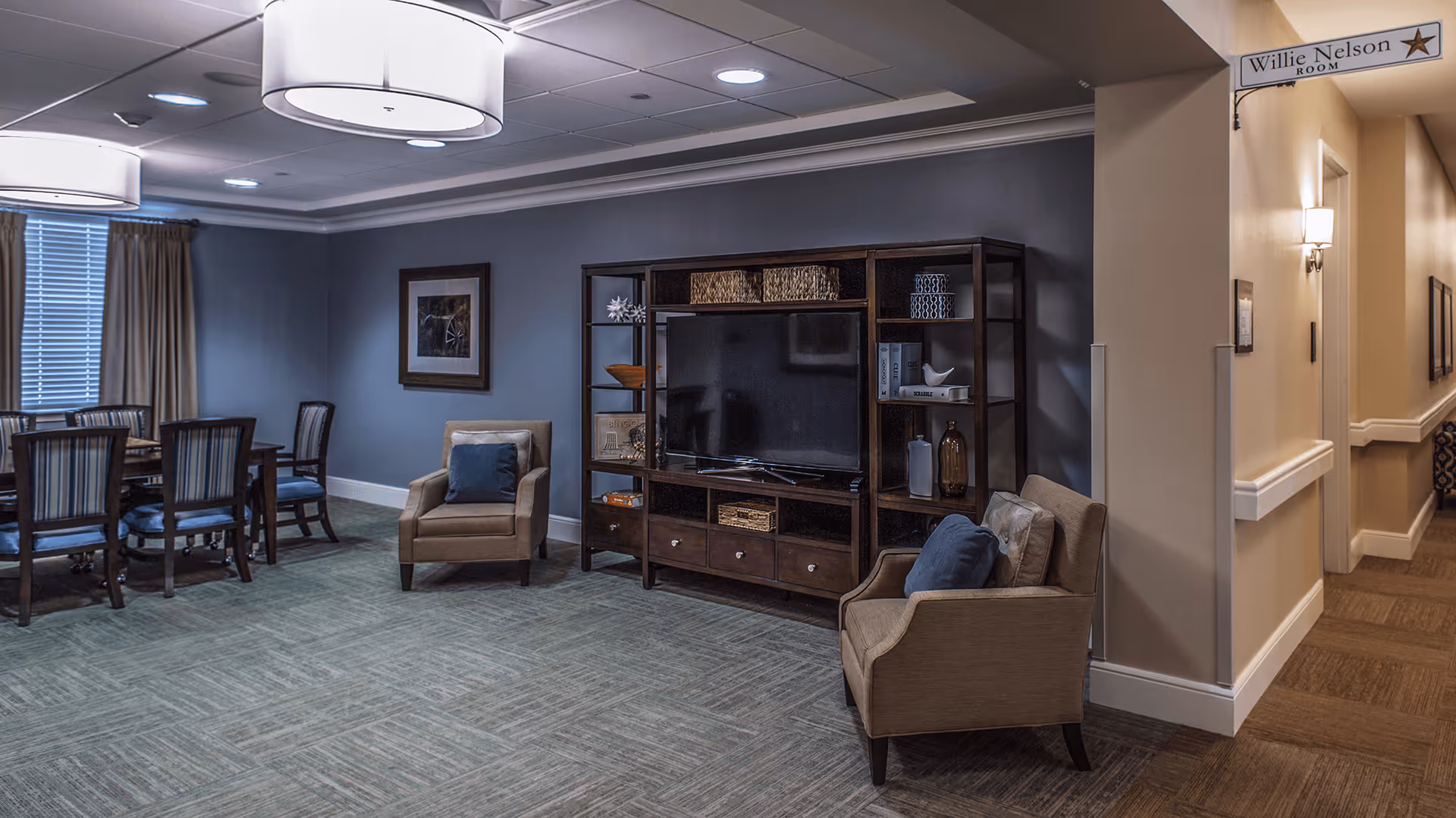 A cozy common area in a senior living facility featuring two beige armchairs with blue cushions, a large dark wood entertainment center with a flat-screen TV, and a dining table with chairs near a window with curtains. The walls are painted in muted tones, and ceiling lights provide soft illumination. A hallway is visible to the right with a sign reading 'Willie Nelson Room.'