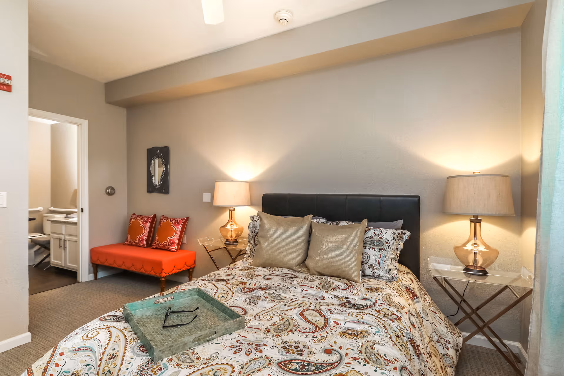 A cozy bedroom with a bed featuring a paisley-patterned comforter and three pillows. On either side of the bed are glass-top nightstands with matching lamps. To the left, there is a bright orange bench with two decorative pillows. An open doorway leads to a bathroom with white cabinetry and a chair. The walls are painted light gray and the room is softly lit.