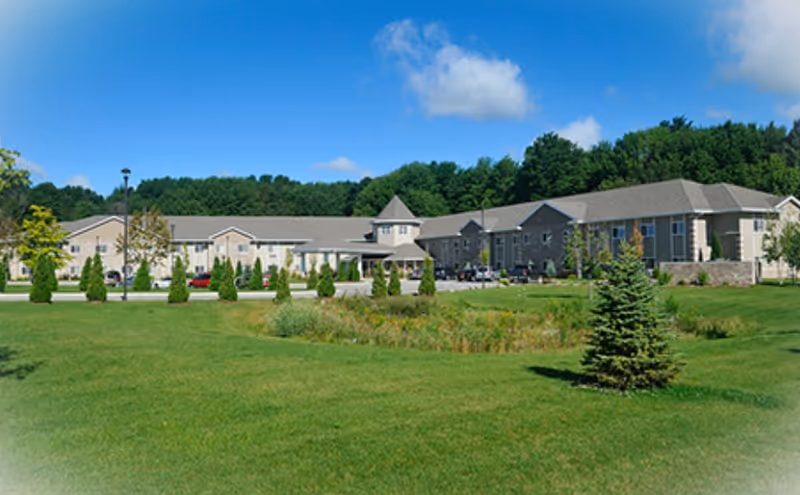 Front exterior of a low-rise assisted living building with a landscaped lawn, driveway, and trees under a blue sky.