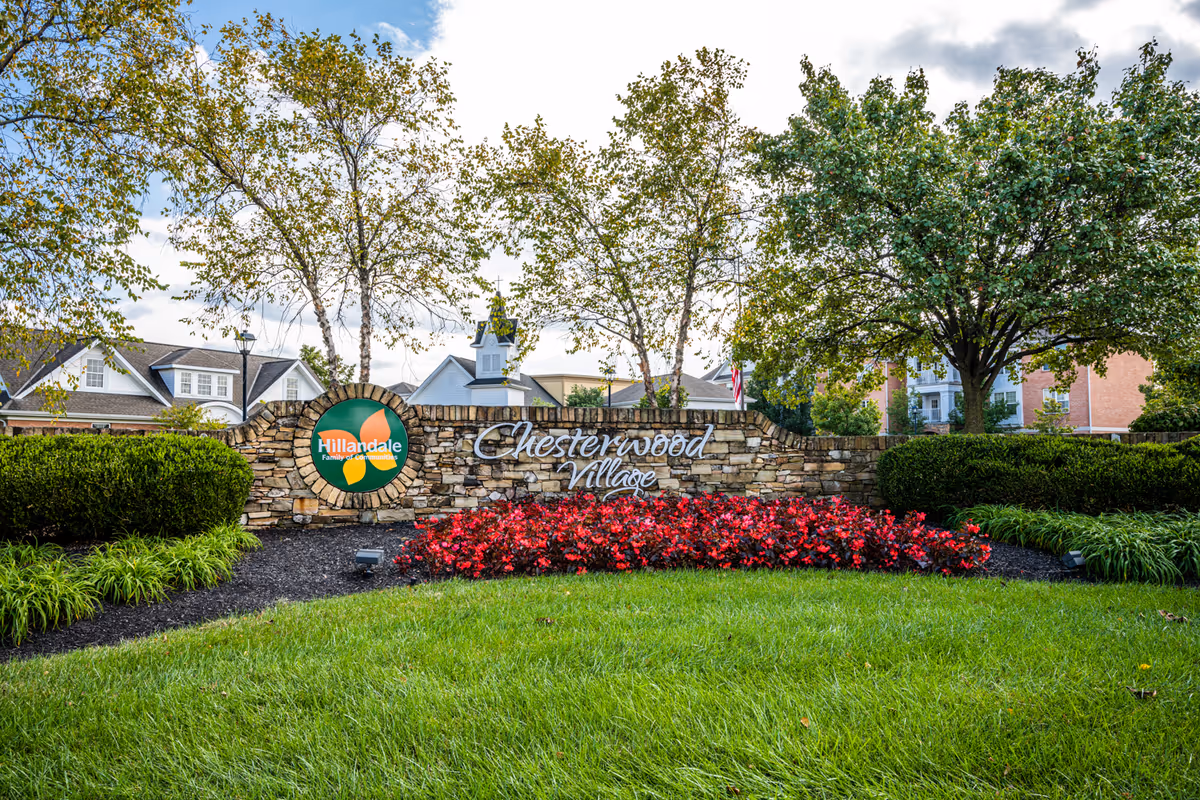 Stone sign for Chesterwood Village with a Hillandale Family of Companies logo, surrounded by green grass, red flowers, bushes, and trees, with residential buildings in the background under a partly cloudy sky.