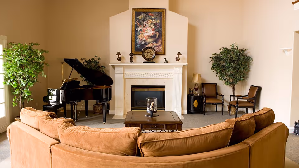 A cozy living room with a large curved brown sofa facing a white fireplace. Above the fireplace is a framed floral painting and decorative items on the mantel. To the left of the fireplace is a black grand piano with a plant behind it. To the right are two wooden chairs with cushions, a small round table with a lamp, and a large potted plant. The room has beige walls and carpeted floor.