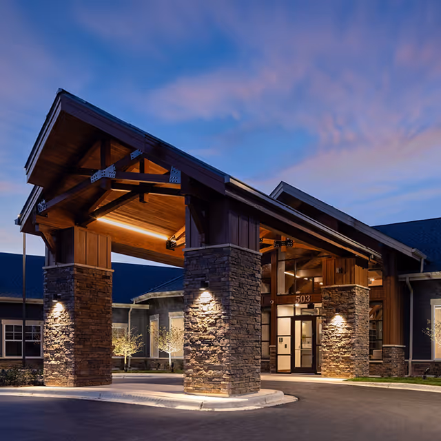 Exterior view of a senior living facility entrance at dusk with a large covered driveway supported by stone pillars and warm lighting under a wooden roof structure.