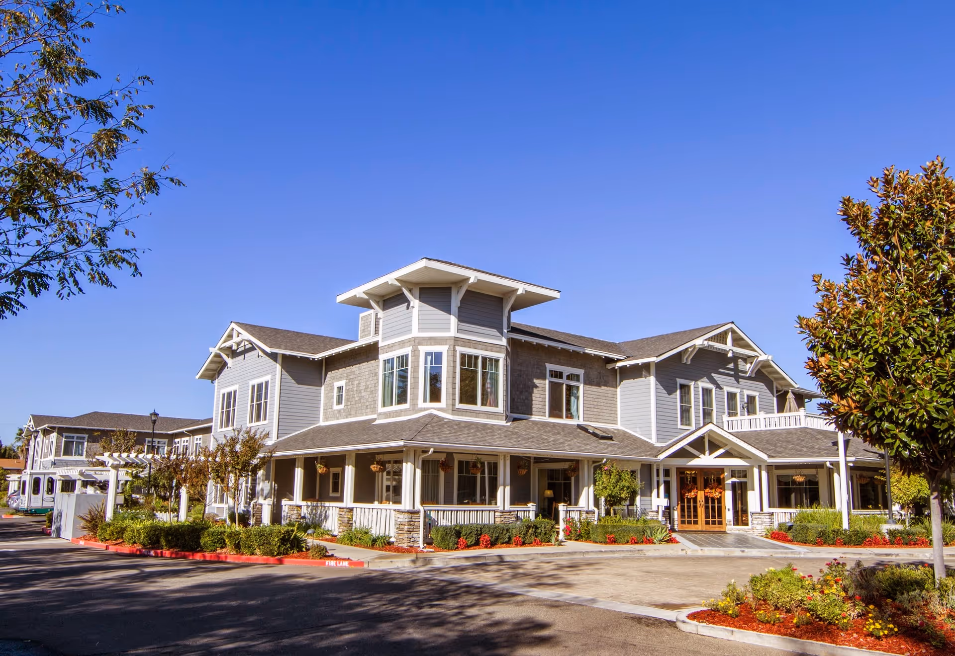 Front exterior of a two-story senior living building with a wraparound porch, landscaped beds, and a clear blue sky.