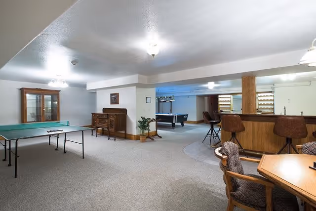 A spacious recreational room featuring a ping pong table on the left, a wooden cabinet and sideboard against the wall, a pool table in the background, and a bar area with three bar stools on the right. There is also a wooden table with cushioned chairs in the foreground. The room has carpeted floors and white walls with ceiling lights.