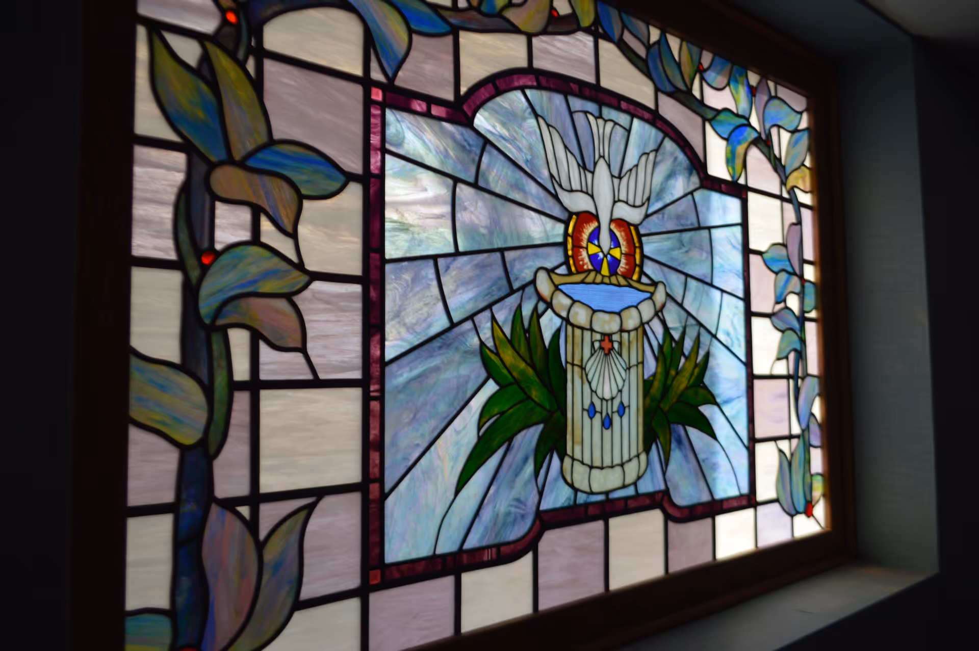 A stained glass window featuring a central design of a white dove above a decorative pedestal with green leaves on either side, surrounded by a border of purple and blue floral patterns.