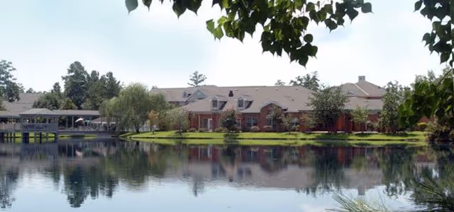 A large single-story brick building with a gray roof situated beside a calm pond, surrounded by trees and greenery. The building is reflected in the water, and there is a covered walkway extending over the pond on the left side.