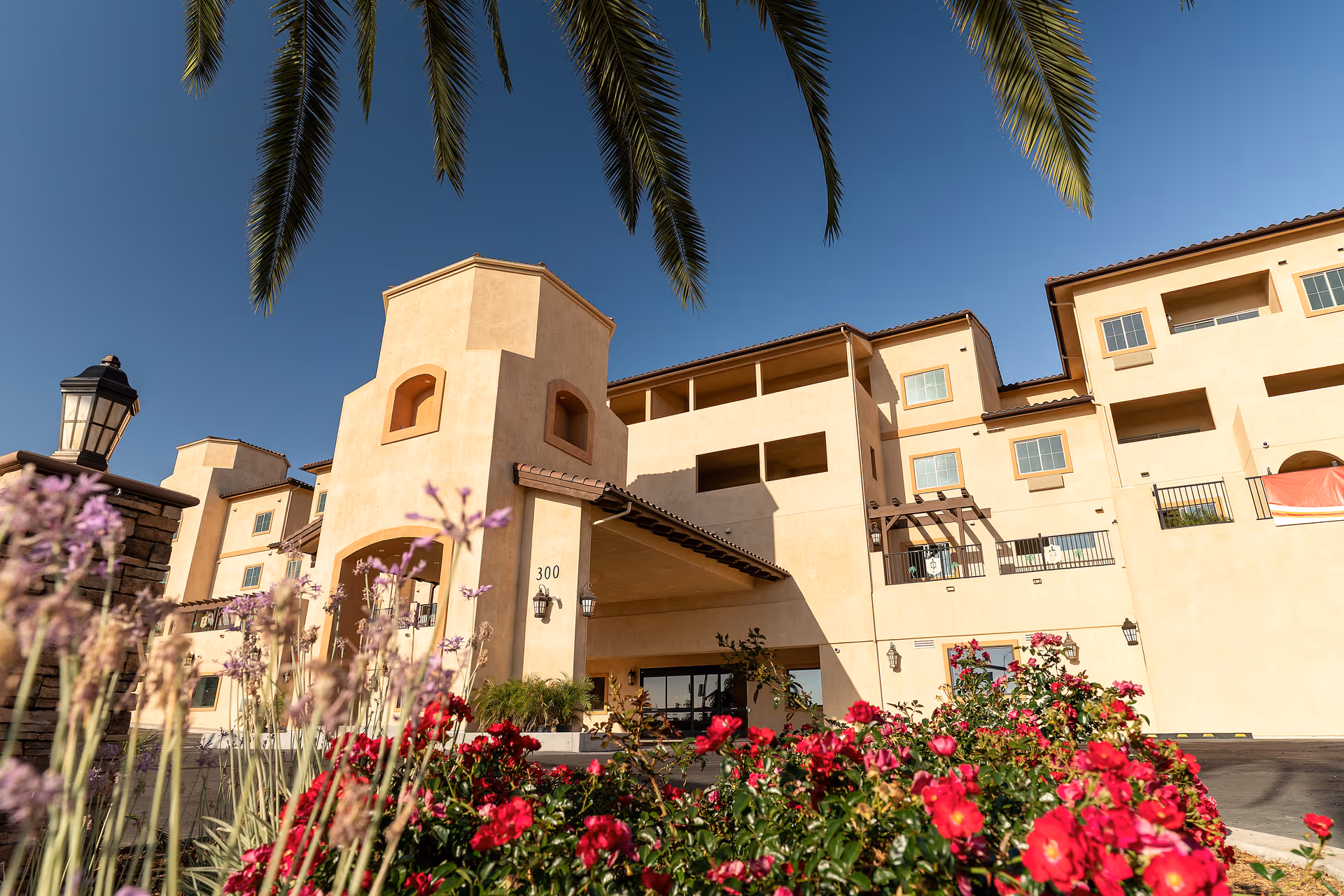 Exterior view of a multi-story senior living facility building with beige stucco walls and a tiled roof, surrounded by vibrant red and purple flowers and palm tree leaves hanging from above, under a clear blue sky.