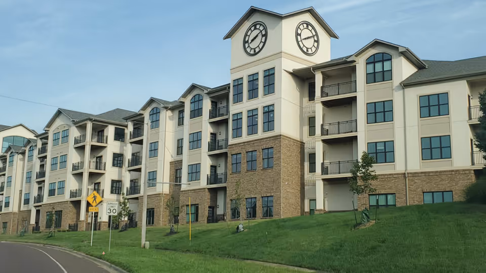 A multi-story senior living building with a central clock tower, balconies, and a grassy lawn in front.
