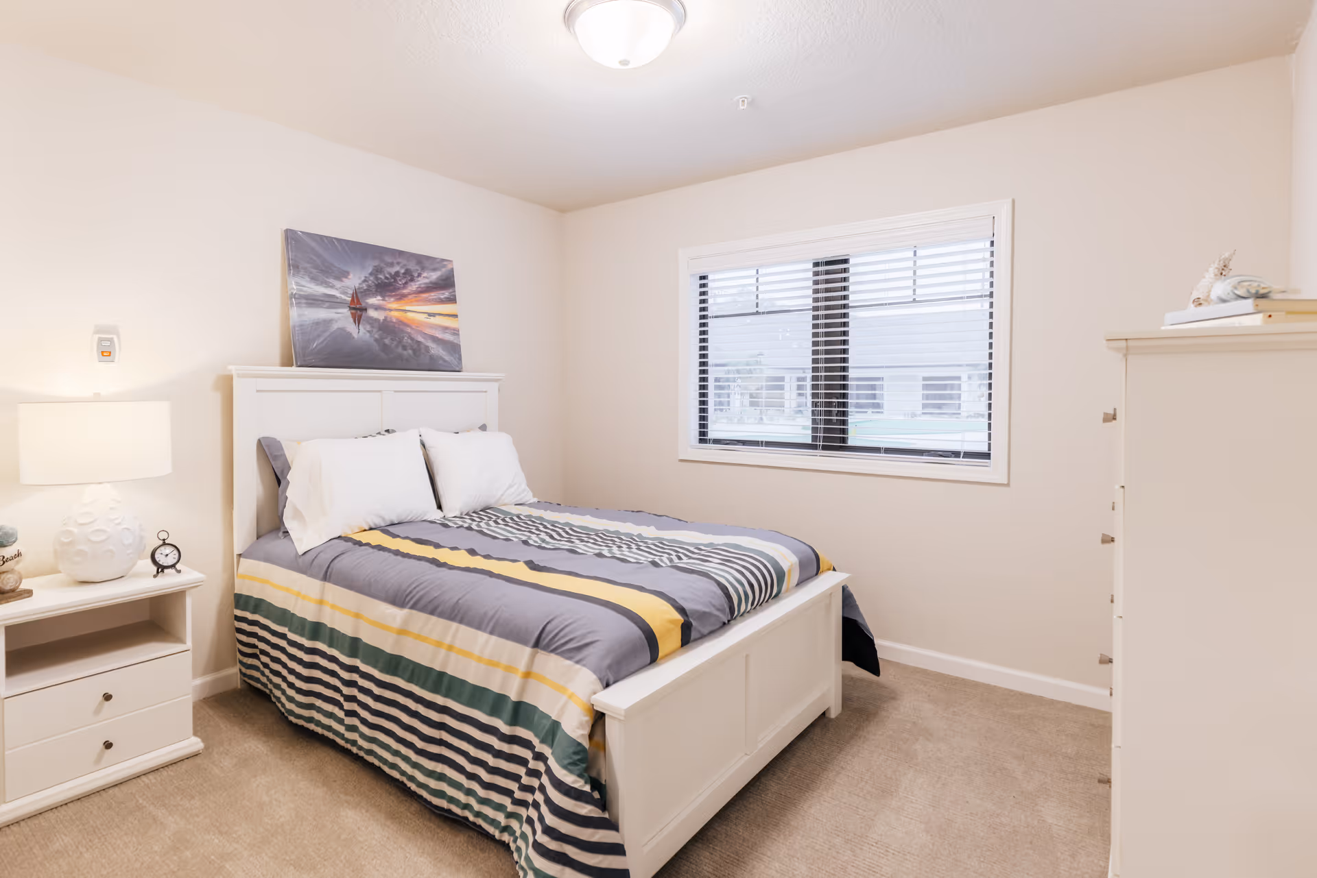 A bright bedroom with a white wooden bed frame and a striped bedspread in shades of gray, yellow, and green. There are two white pillows on the bed. A nightstand with a white lamp and a small clock is positioned to the left of the bed. A colorful painting of a sailboat at sunset is hung above the bed. A window with white blinds is on the right wall, and a white dresser with books and decorative items is partially visible on the right side of the room. The walls are light-colored, and the floor is carpeted.