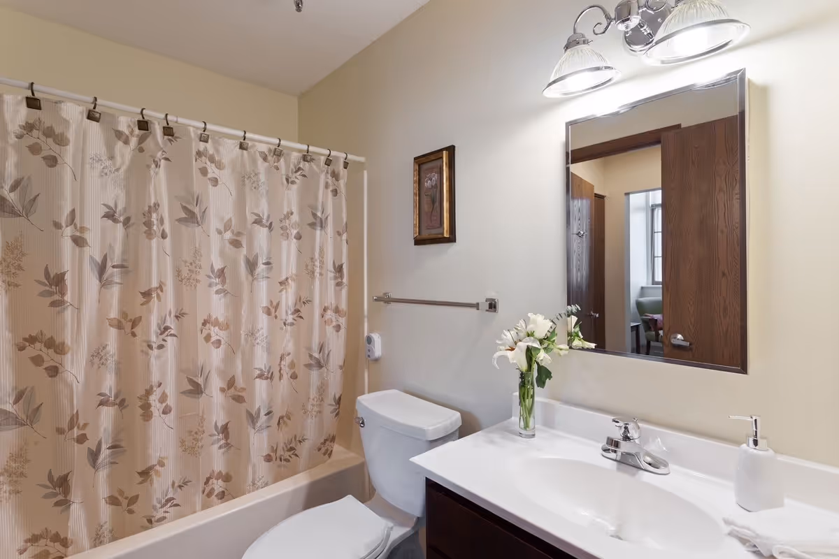 A clean bathroom with a white toilet and a bathtub with a floral-patterned shower curtain. There is a white sink with a silver faucet, a soap dispenser, and a vase with white flowers on the countertop. Above the sink is a rectangular mirror with a wooden frame, and above the mirror are two light fixtures with glass shades. A small framed picture hangs on the wall next to the mirror.