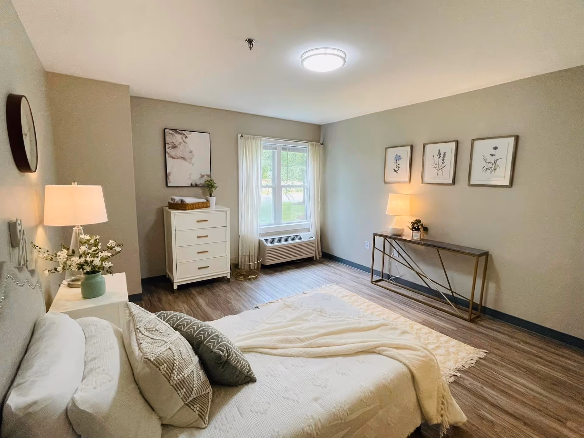 A bright and cozy bedroom in an assisted living facility featuring a neatly made bed with white bedding and decorative pillows. There is a nightstand with a lamp and flowers beside the bed. Across the room, a white chest of drawers stands near a window with sheer curtains, and a console table with a lamp and framed botanical prints hangs on the wall above it. The room has wood flooring and neutral-colored walls.