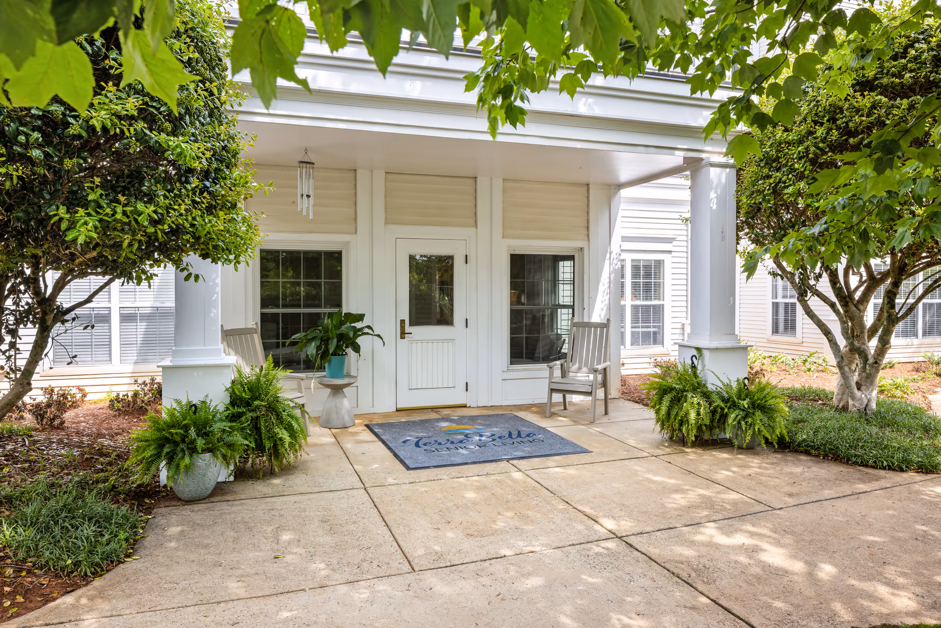 Covered entrance of a white senior living building with a door, two chairs, potted ferns, and a TerraBella welcome mat.