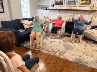 Three elderly individuals seated in a living room, engaging in a group exercise or stretching activity with their arms raised. A woman is seated facing them, possibly leading the activity. The room features a fireplace with decorative items on the mantel, a large rug, and comfortable seating including a couch and chairs.