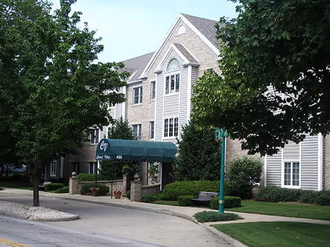 Three-story brick and siding senior living building with a green awning over the entrance, trees, and a driveway.