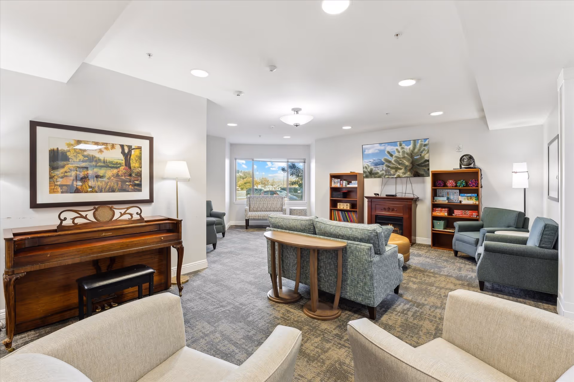 A bright and spacious living room area in a senior living facility with multiple seating options including armchairs and sofas arranged around a small wooden table. There is a wooden piano with a bench on the left side, a large framed landscape painting above it, and a floor lamp nearby. On the far wall, a flat-screen TV is mounted above a fireplace, flanked by two bookshelves filled with books and games. A large window lets in natural light, and the room has a neutral color palette with carpeted flooring.