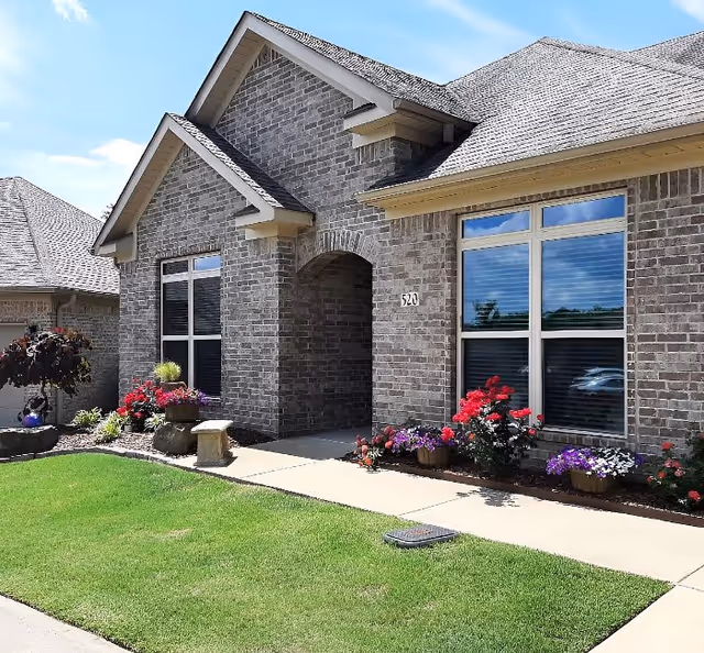 Exterior view of a brick building with a peaked roof, large windows, and a small garden with colorful flowers along the sidewalk leading to the entrance.