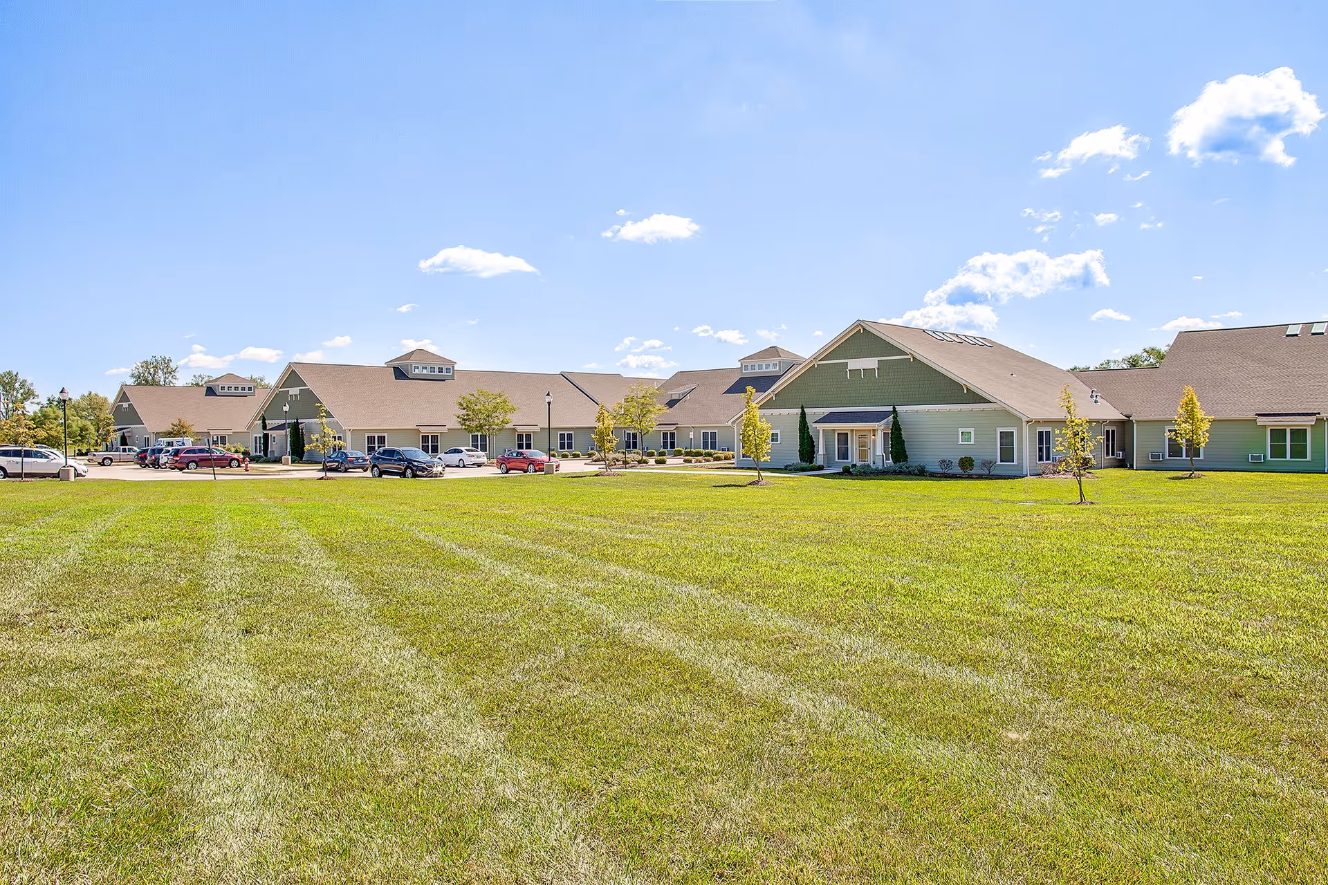 Wide view of a senior living facility building with a large green lawn in the foreground under a clear blue sky with some clouds. Several cars are parked near the building, which has a beige and green exterior with multiple roof sections and small trees planted around.