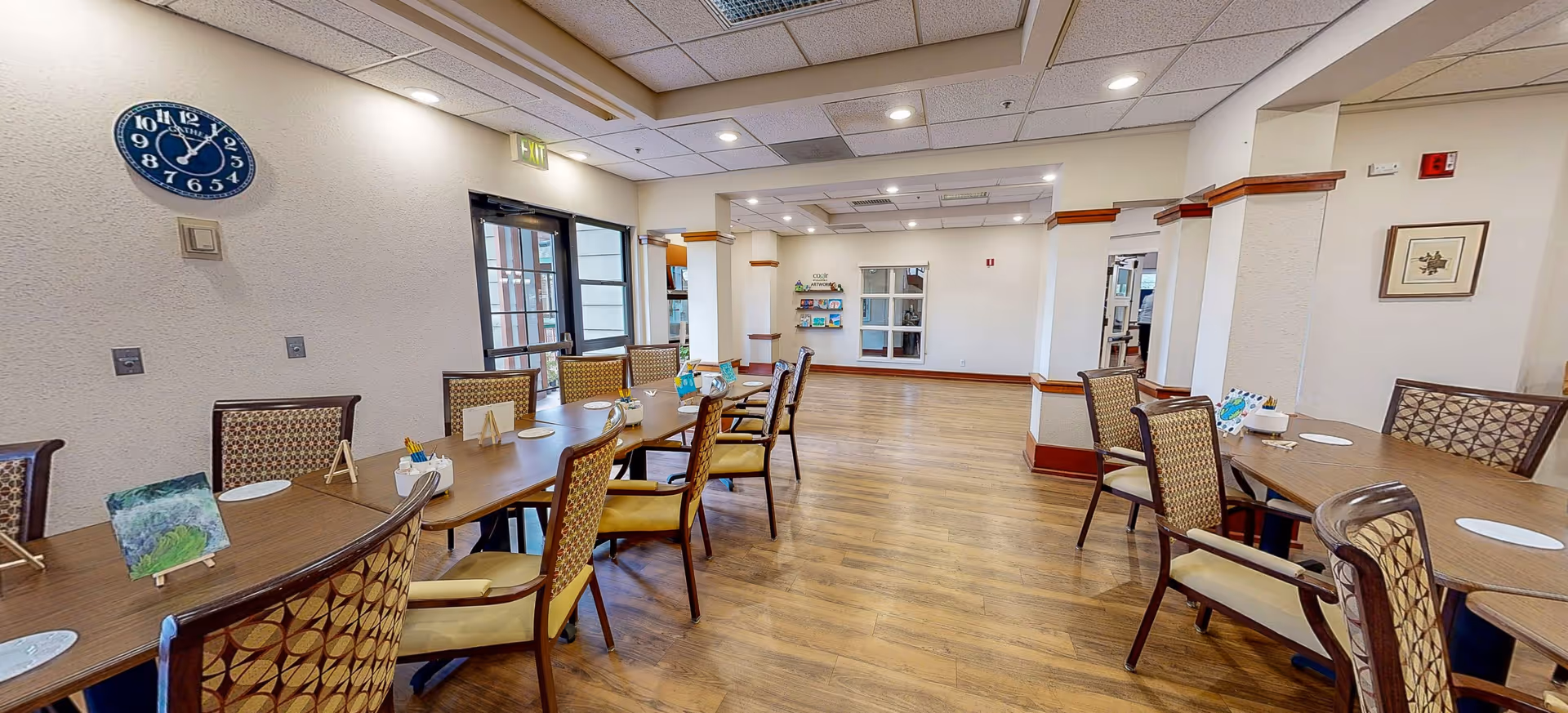 A spacious senior living facility common area with wooden flooring and several tables and chairs arranged for group activities or dining. The walls are light-colored with a blue clock showing 11:55, and there are small paintings and decorations on the tables. The room is well-lit with ceiling lights and has large windows and doors allowing natural light.