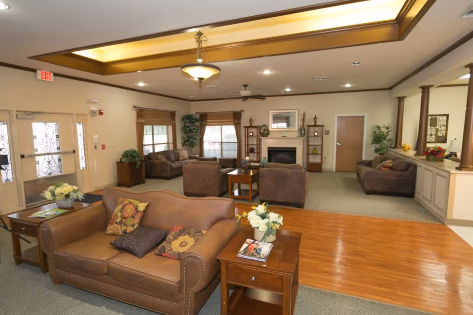 A spacious living room area in a senior living facility with multiple brown sofas and armchairs arranged around wooden coffee tables. The room features a fireplace with decorative shelves on either side, large windows with blinds, potted plants, and a ceiling fan. The floor is a combination of carpet and wood, and there is a decorative light fixture on the ceiling.