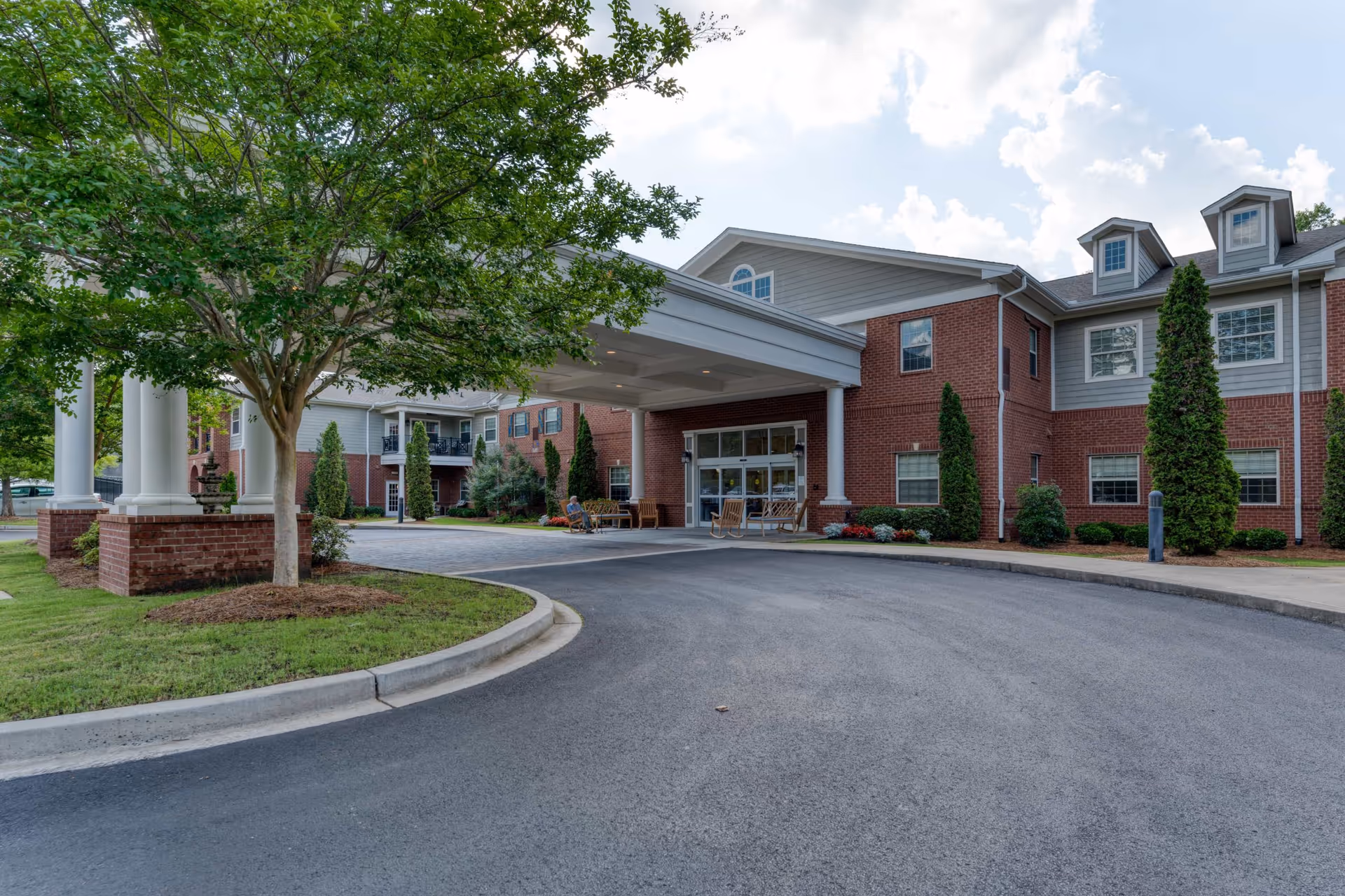 Entrance of Camellia Walk of Evans senior living facility showing a covered drop-off area with benches and rocking chairs, surrounded by a well-maintained landscape with trees and shrubs, under a partly cloudy sky.