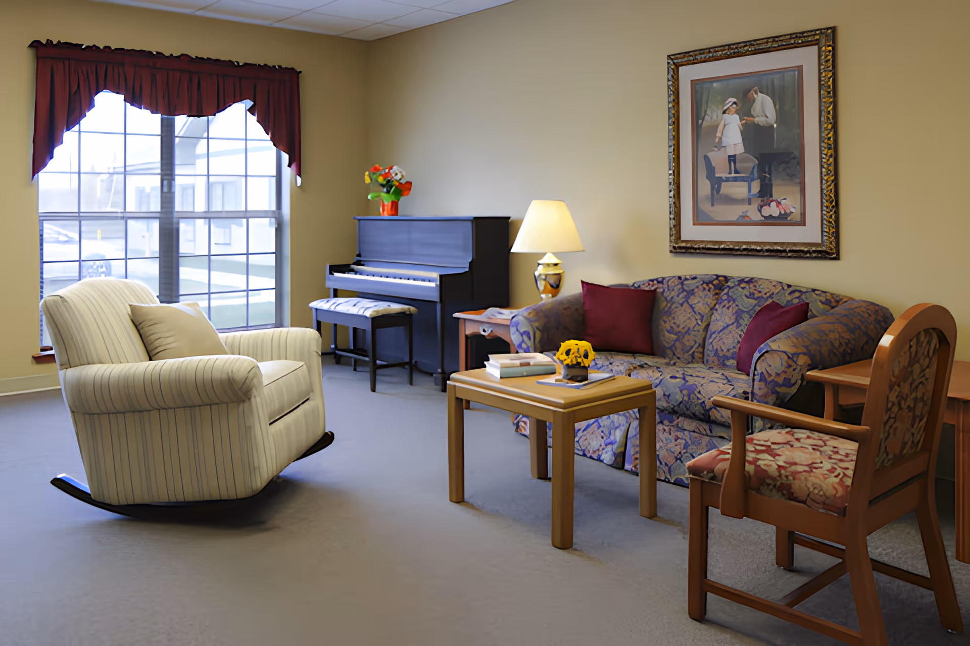 A cozy living room with a striped rocking chair, a floral-patterned sofa with red cushions, a wooden coffee table with books and a small flower arrangement, a wooden armchair with floral upholstery, a piano with a cushioned bench, a table lamp, and a framed painting on the wall. A large window with red curtains lets in natural light.