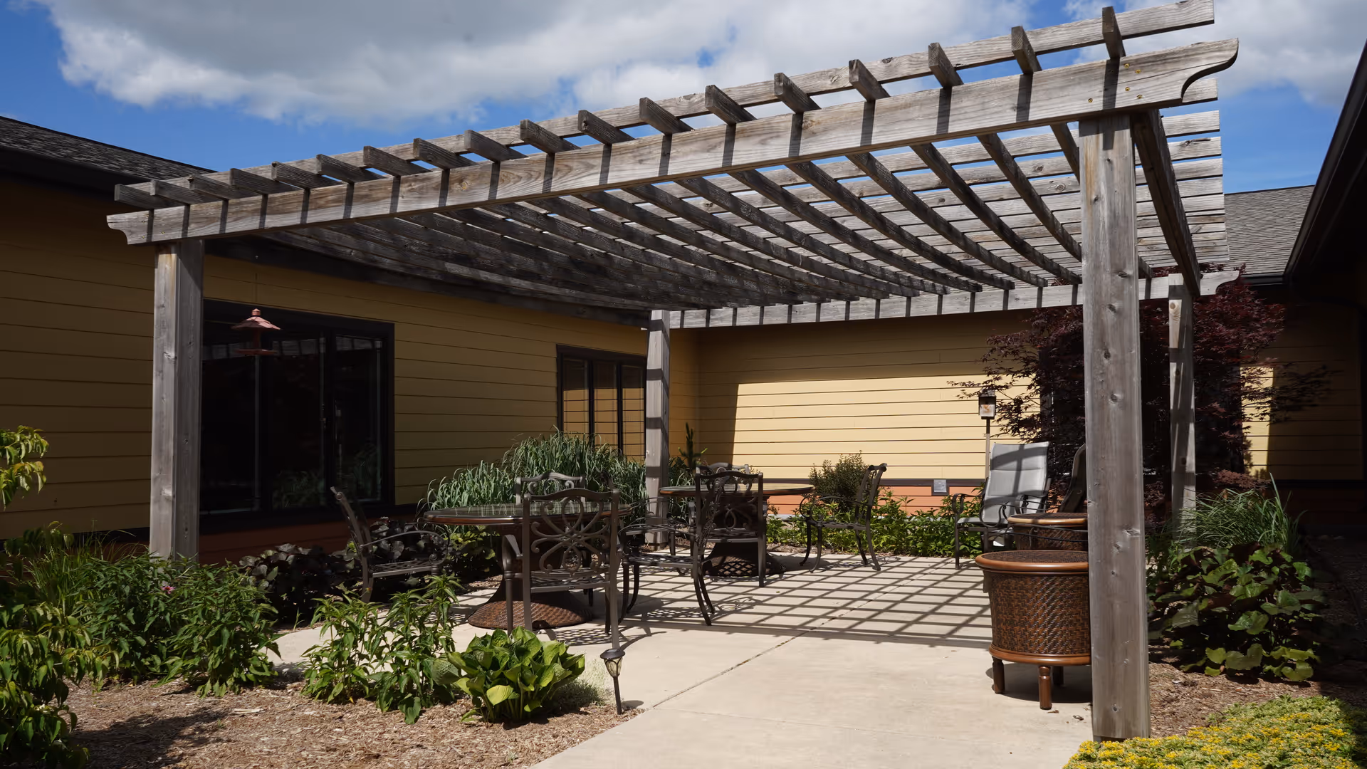 Outdoor patio area with a wooden pergola casting shadows on the concrete floor, surrounded by garden plants and shrubs. There are metal chairs and tables arranged under the pergola, with a yellow building wall in the background under a partly cloudy sky.