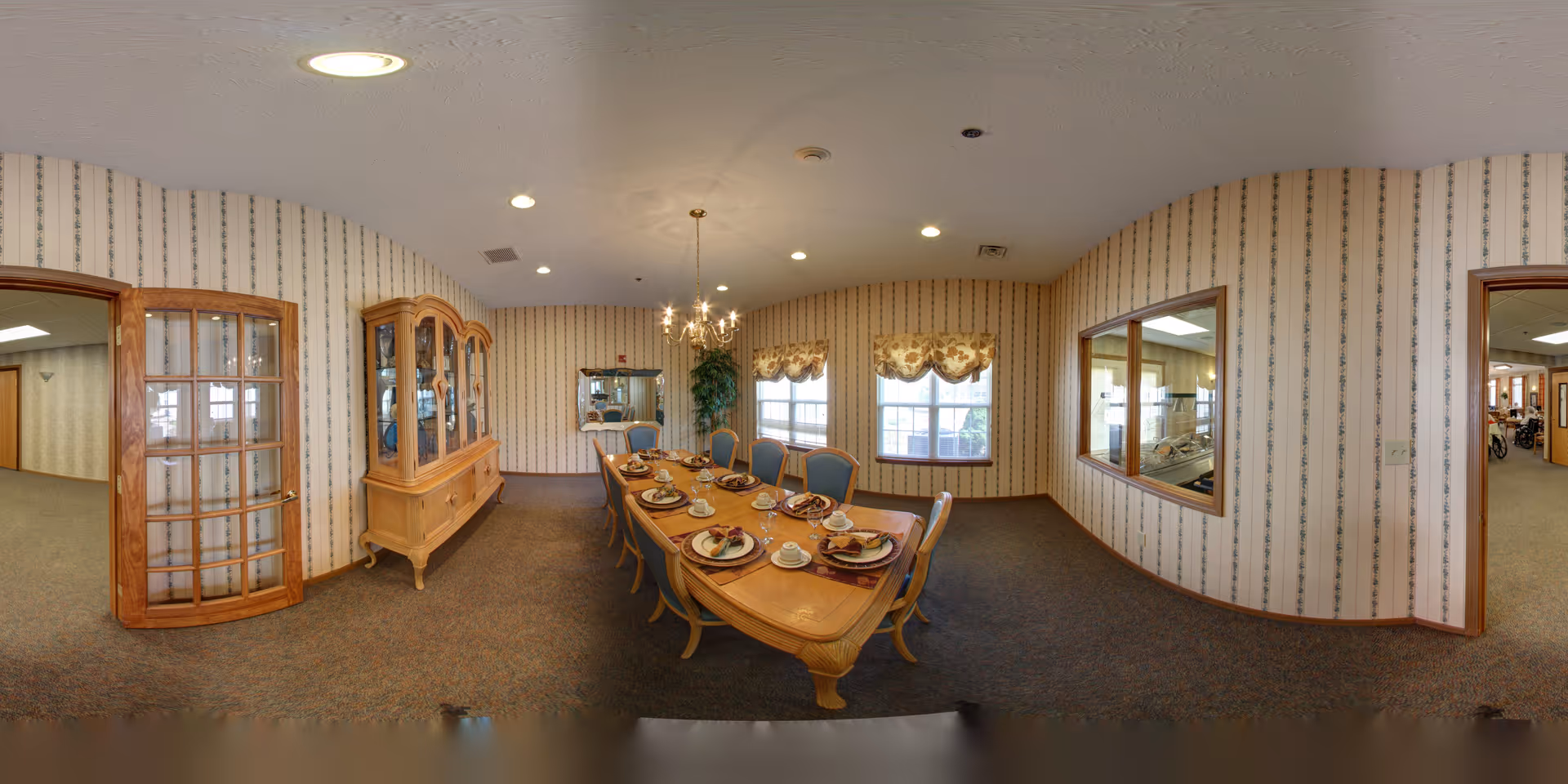 A dining room with a long wooden table set for eight people, featuring plates, cups, and napkins. The room has patterned wallpaper, carpeted floor, a chandelier hanging above the table, a wooden china cabinet, and windows with floral valances. There are two doorways and a large interior window showing an adjacent room.