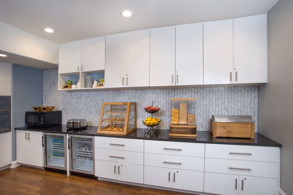 A modern kitchen area with white cabinets, a black countertop, and a herringbone patterned backsplash. On the counter, there is a microwave, a toaster, a display case with pastries, a two-tier fruit stand with apples and lemons, a cereal dispenser, and a wooden bread box. The floor is wooden, and the space is well-lit with recessed ceiling lights.