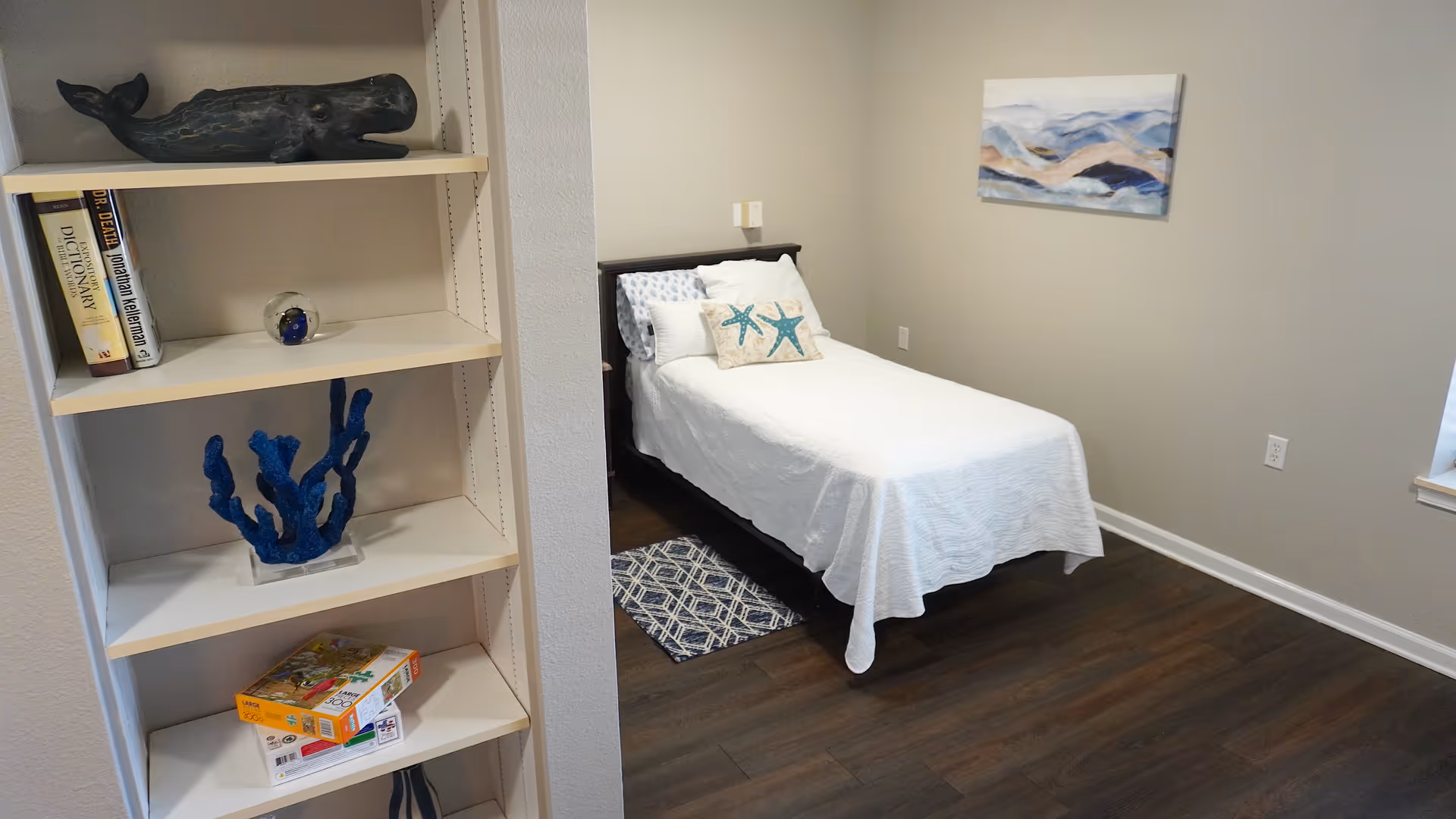 A small bedroom with a single bed covered in white bedding and two starfish-themed pillows. There is a blue patterned rug partially under the bed, a painting of abstract mountains on the wall, and a window on the right side. On the left, a white bookshelf holds books, a blue coral decoration, a wooden whale sculpture, and some board games.
