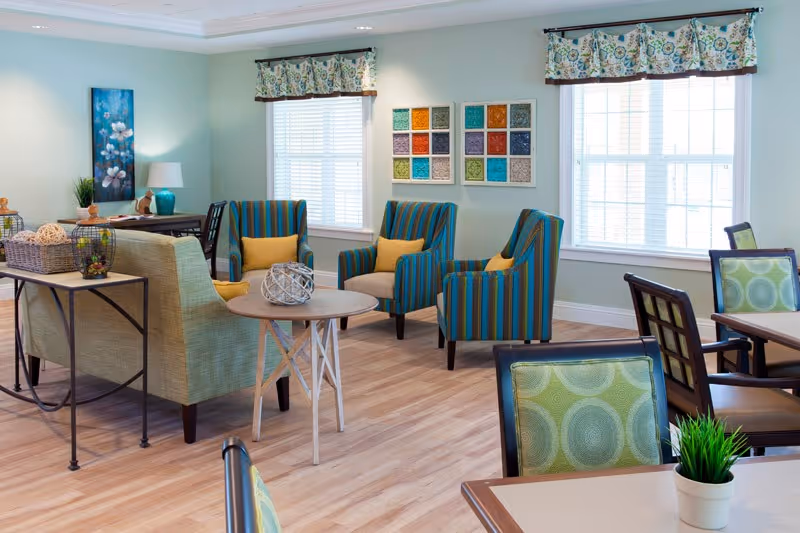A bright and cozy living room area in a senior living facility with light blue walls, two large windows with floral valances, three striped armchairs with yellow cushions, a green sofa, a round wooden side table, and several dining tables with green patterned chairs. The room is decorated with colorful wall art and small plants.