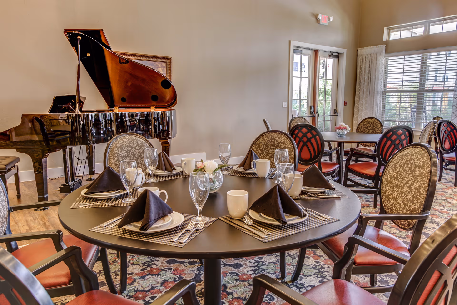 A dining room with round tables set for a meal, featuring black napkins, white plates, cups, and glasses. The room has patterned chairs with red and beige upholstery, a floral carpet, large windows with blinds, and a grand piano in the corner.