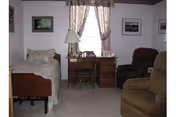 Furnished bedroom with a single bed, a desk and chair under a curtained window, two armchairs, and framed pictures on the walls.