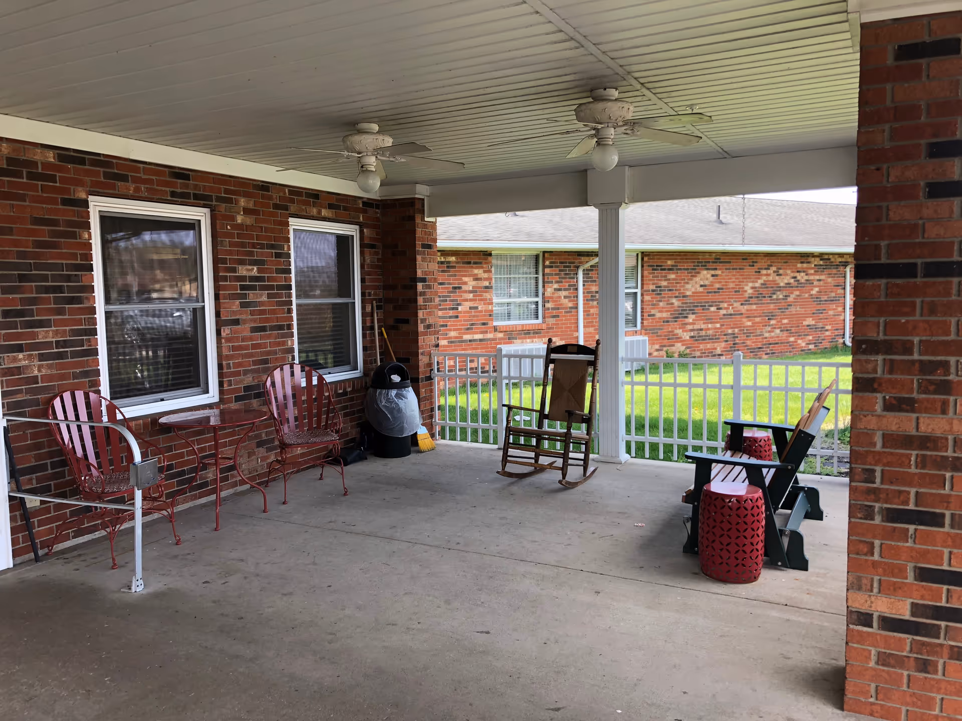 Covered outdoor patio area with a concrete floor and white ceiling featuring two ceiling fans. There are two red metal chairs with a small matching table against a brick wall with two windows. A black trash can and broom are placed near the corner. On the right side, there is a wooden rocking chair and two dark green Adirondack chairs with two red decorative stools. A white picket fence and green grass are visible beyond the patio.