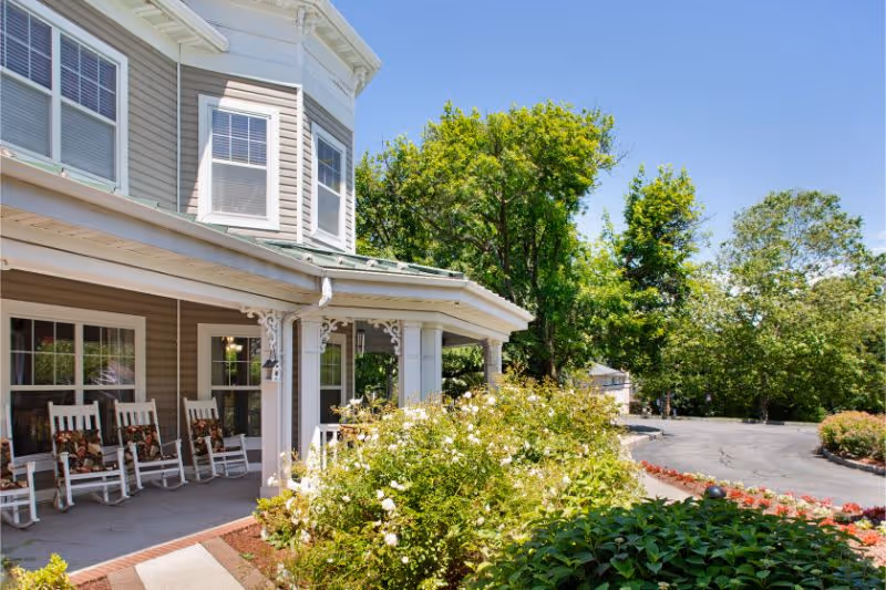 Exterior view of a senior living facility with a covered porch featuring several white rocking chairs with floral cushions. The building has beige siding and white trim, surrounded by green trees and flowering bushes under a clear blue sky.