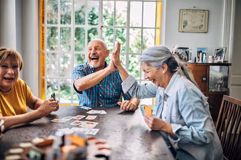 Three elderly people sitting around a table playing cards, two of them giving each other a high five and smiling joyfully in a bright room with large windows.