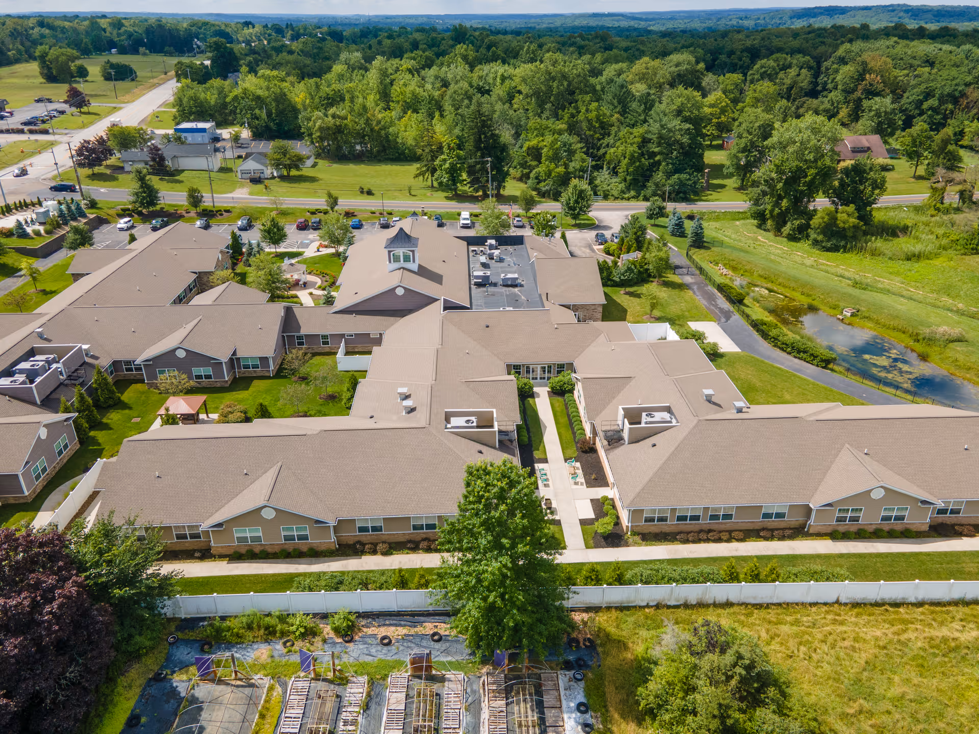 Aerial view of a single-story senior living complex with connected beige buildings, lawns, parking and a nearby pond.