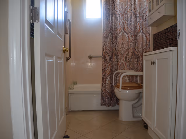 View of a bathroom with a partially open door, a bathtub with a patterned shower curtain, a toilet with armrests, a cabinet with a sink, and a small window letting in natural light.
