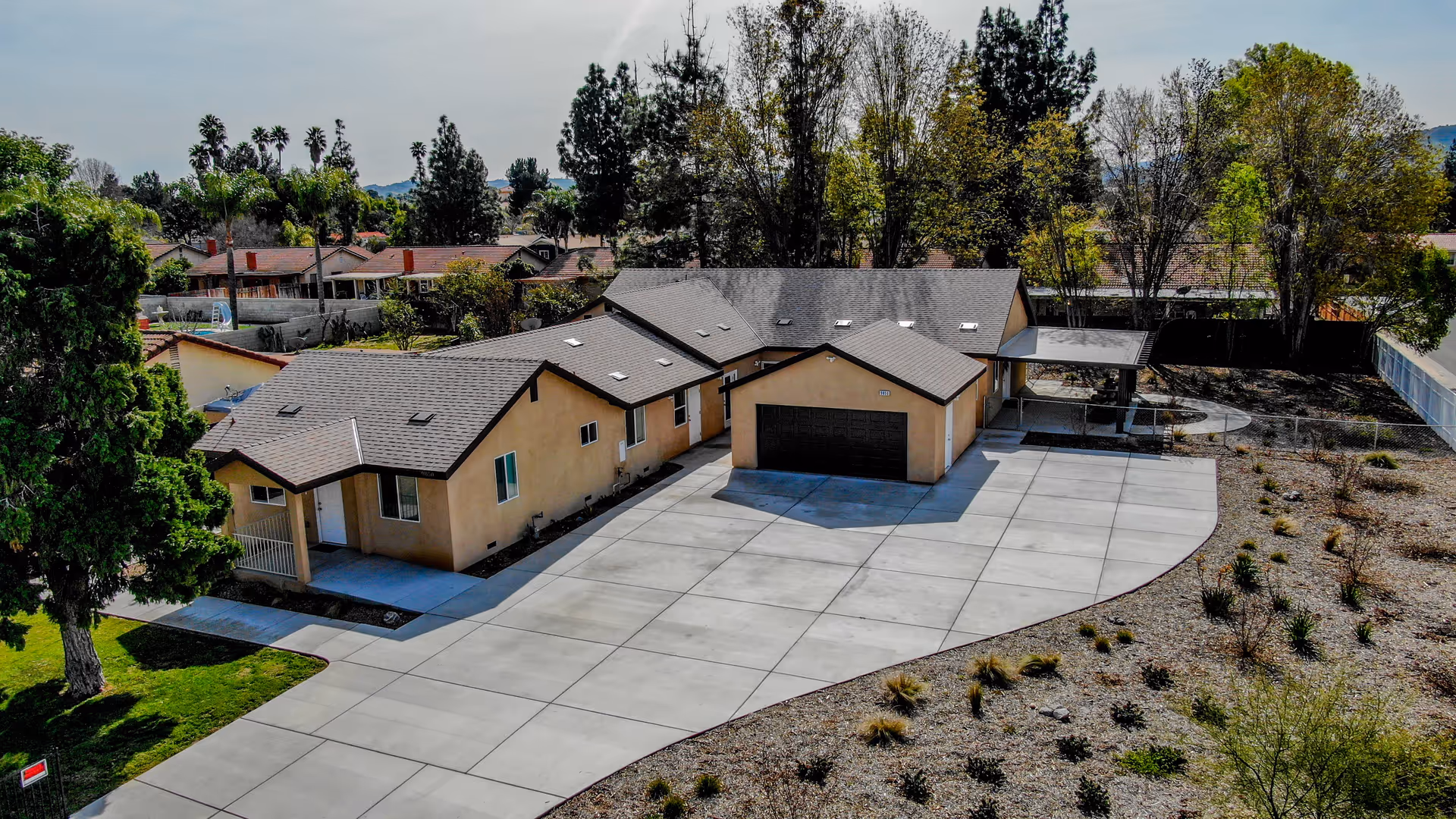 Aerial view of a single-story residential building with a large concrete driveway, surrounded by trees and landscaping. The building has a beige exterior with a dark roof and a detached garage. The surrounding area includes other houses and greenery.